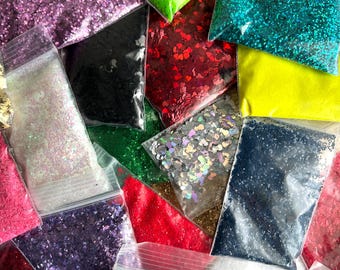 May include: An assortment of glitter in various colors, including pink, green, blue, black, and silver, packaged in clear plastic bags. Some glitter has a heart shape. The glitter is used for crafts and decorations.