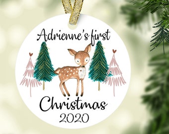 May include: White round Christmas ornament with a deer and trees. The ornament says "Adrienne's first Christmas 2020".