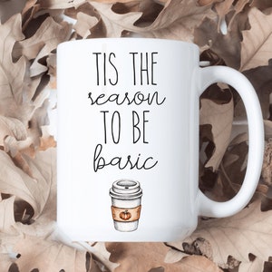 Peut inclure: Mug en céramique blanche avec l'inscription "Tis the season to be basic" en écriture noire. Un dessin d'une tasse à café avec un motif de citrouille épicée est en dessous du texte. Le mug est posé sur un fond de feuilles brunes séchées.