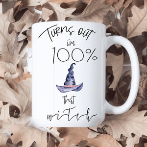 May include: White ceramic mug with the text "Turns out I'm 100% that witch" and a watercolor illustration of a witch's hat. The mug is set against a background of dried brown leaves.