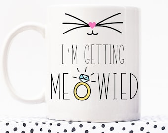 May include: White ceramic mug with a black cat face and pink heart nose. The mug says "I'm getting MEOWIED" with a yellow ring and blue diamond.