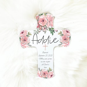 May include: A white wooden cross decorated with pink roses and green leaves. The name "Addie" is written in black script. The cross includes the text "Baptized September 20, 2020" and a religious quote.