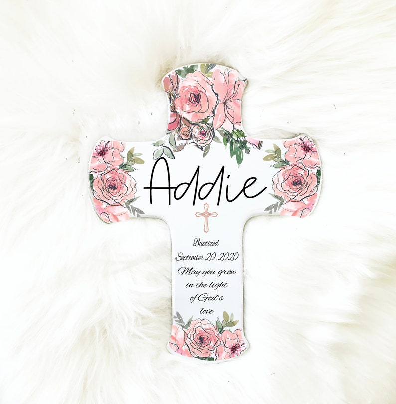 May include: A white wooden cross decorated with pink roses and green leaves. The name "Addie" is written in black script. The cross includes the text "Baptized September 20, 2020" and a religious quote.