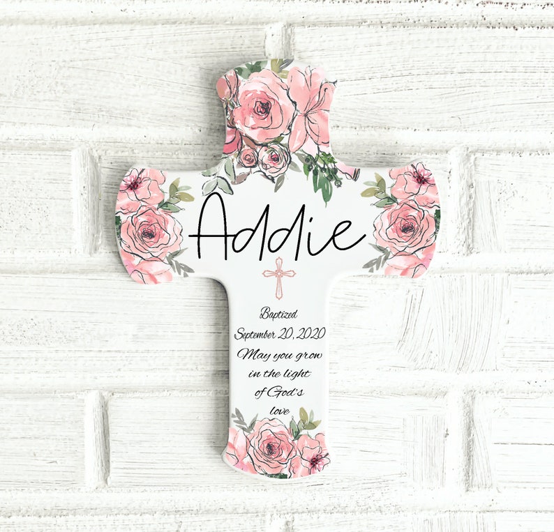 May include: White wooden cross with pink floral accents and the name "Addie" in black script. The cross includes the text "Baptized September 20, 2020" and a religious quote. The cross is a religious item.