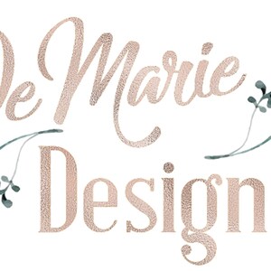 May include: The logo features the text "De Marie Designs" in a textured, rose gold font. Delicate watercolor floral elements in pink and green accent the text, set against a white background.