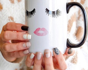 May include: White ceramic mug with a black handle. The mug has a pink watercolor lip design and black eyelashes on the front.