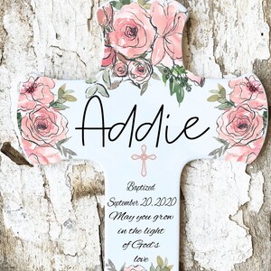 May include: A white cross-shaped plaque with watercolor-style pink roses and green leaves. The name "Addie" is written in black script. The plaque includes the text "Baptized September 20, 2020 May you grow in the light of God's love."