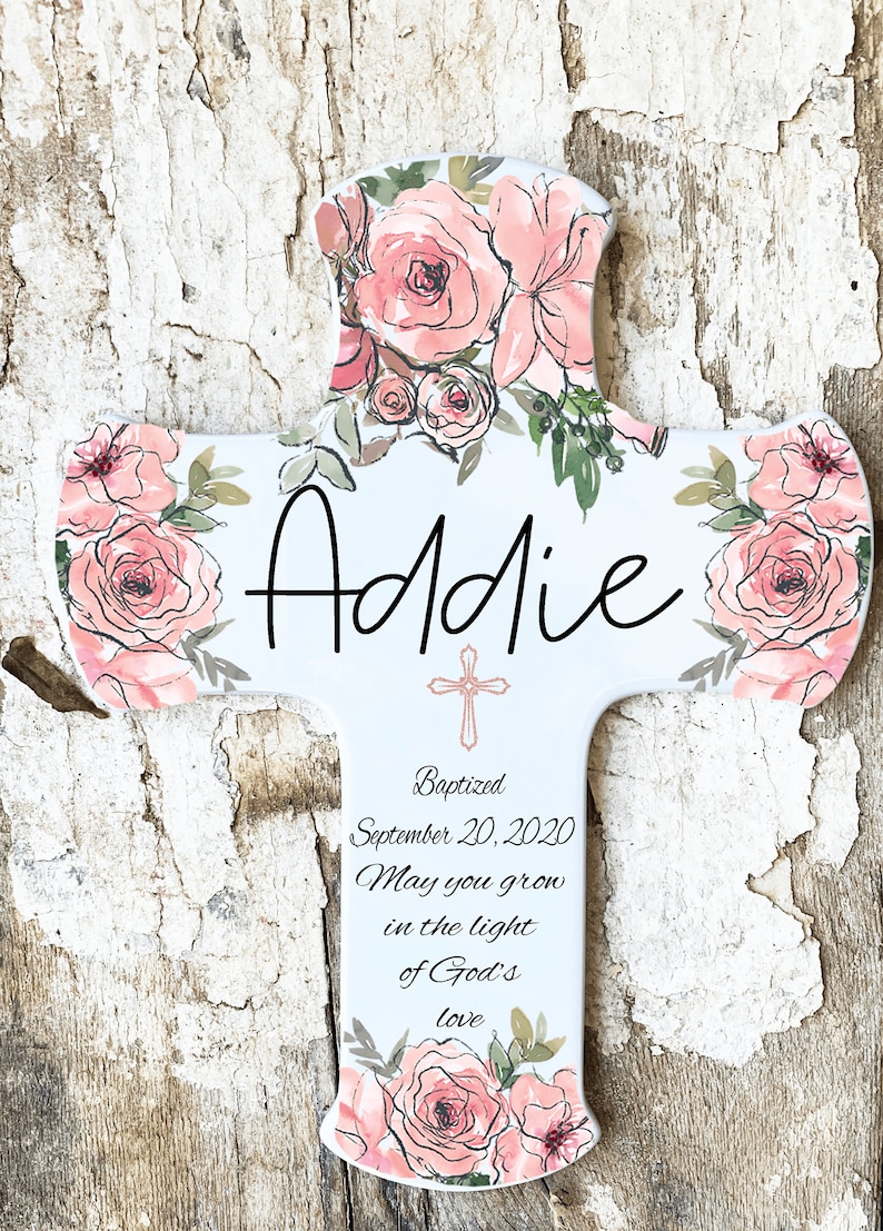 May include: A white cross-shaped plaque with watercolor-style pink roses and green leaves. The name "Addie" is written in black script. The plaque includes the text "Baptized September 20, 2020 May you grow in the light of God's love."