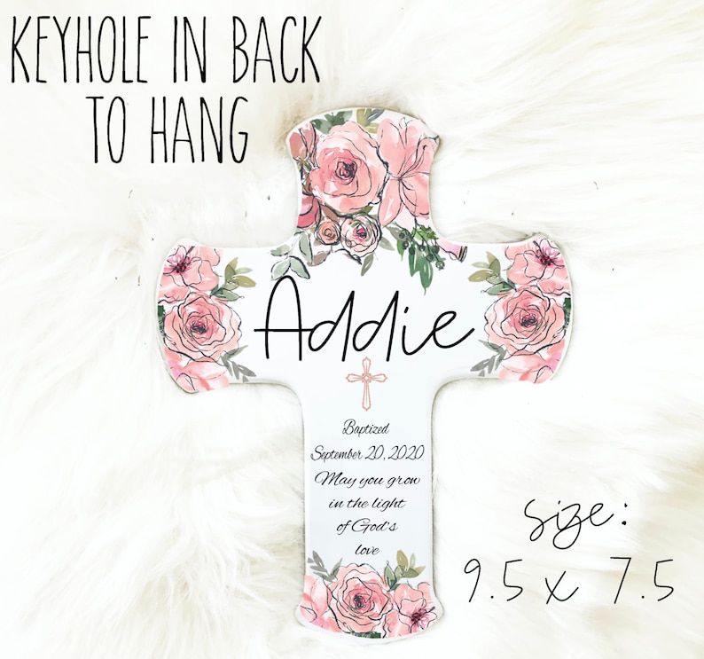 May include: A white wooden cross decorated with pink watercolor roses and green leaves. The name "Addie" is written in black script. The cross is 9.5 x 7.5 inches and includes the text "Baptized September 20, 2020" and "May you grow in the light of God's love."