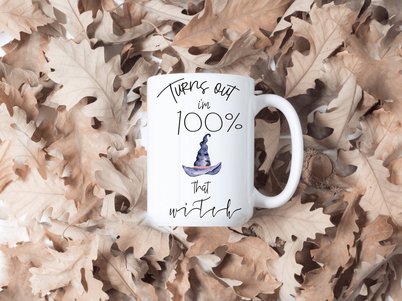 May include: White ceramic mug with the text "Turns out I'm 100% that witch" and a watercolor illustration of a witch's hat. The mug is set against a background of dried brown leaves.