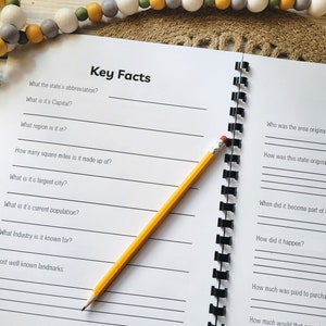 May include: A blank notebook with a spiral binding and lined pages. The notebook is open to a page with the title "Key Facts" and a list of questions about a state. The next page is titled "History" and has questions about the history of a state. A yellow pencil is lying on the notebook.