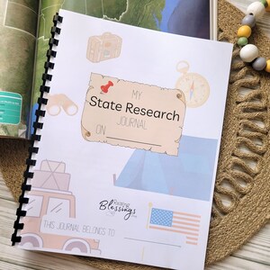 May include: A white journal with a colourful illustration of a car, a tent, binoculars, a suitcase, and a compass. The text "My State Research Journal" is on the cover. The journal is bound with a black coil.