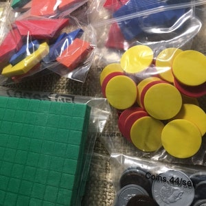 May include: Assorted colorful educational manipulatives, including geometric shapes, counters, and a green base-ten block, packaged in clear plastic bags. The shapes are red, blue, yellow, and green. The coins are silver and brown.