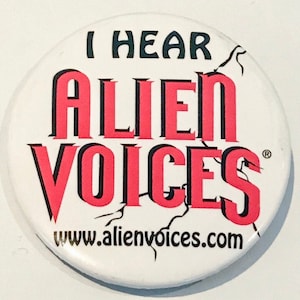 May include: White button with black text that reads "I HEAR ALIEN VOICES" in a bold, stylized font. The text "ALIEN VOICES" is in pink. Below the text is a website address in black: www.alienvoices.com. There is a small registered trademark symbol in the upper right corner.