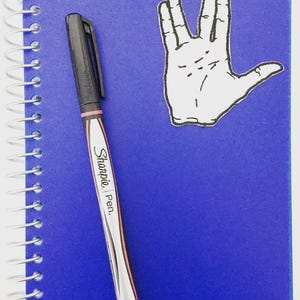 May include: A blue notebook with a spiral binding, a black Sharpie pen, and a white sticker of the Vulcan salute hand gesture. The notebook has the word "stellar" printed on the bottom right corner.