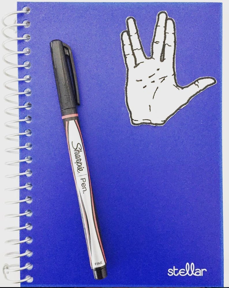 May include: A blue notebook with a spiral binding, a black Sharpie pen, and a white sticker of the Vulcan salute hand gesture. The notebook has the word "stellar" printed on the bottom right corner.
