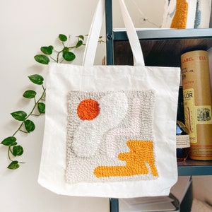 May include: A white canvas tote bag with a hand-stitched design featuring abstract shapes in orange, white, pink, and yellow.