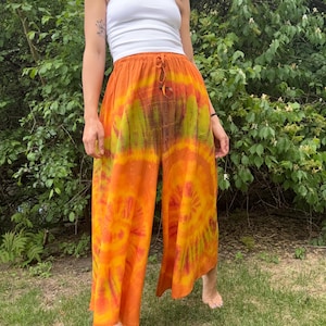 Puede incluir: Un top blanco sin tirantes combinado con pantalones de pernera ancha teñidos con tintes en tonos naranja, amarillo y verde. Los pantalones tienen una cintura con cordón. El atuendo se usa al aire libre con un fondo de hojas verdes.
