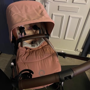 Könnte beinhalten: Ein rosa Kinderwagen mit schwarzem Gestell und braunen Lederakzenten. Der Kinderwagen hat ein rosa Verdeck und einen rosa Fußsack.