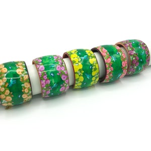 May include: Five colourful wooden bangles with a floral and bird design. The bangles are green, yellow, pink, and purple with white flowers and blue birds.