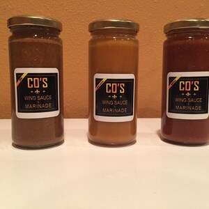 Peut inclure: Trois bouteilles en verre de marinade pour sauce à ailes, étiquetées "CO'S WING SAUCE & MARINADE". Les bouteilles sont brunes, jaunes et rouges.