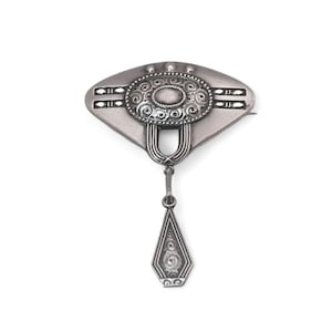 Può includere: Un broche pendente in argento con un design geometrico. Il broche ha un pendente a forma di lacrima con un design circolare e una parte superiore a forma di ventaglio con un design circolare.