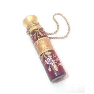 May include: An antique perfume bottle with a cylindrical, deep red glass body. It has gold-coloured metal accents, including a decorative band and cap. The glass is decorated with delicate white and pink floral patterns, and a gold chain is attached.