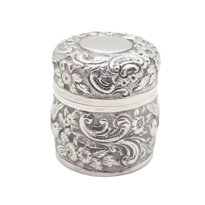 May include: A silver-coloured, cylindrical container with a lid. The container and lid feature detailed floral and scrollwork patterns. The design is symmetrical, with the floral patterns wrapping around the body.