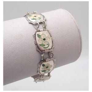 May include: Silver-toned bracelet featuring rectangular links with floral designs. Each link showcases a white flower with green leaves and a black center. The bracelet is displayed on a white surface, highlighting its intricate details.