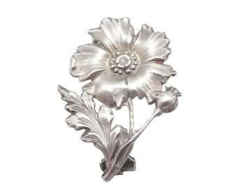 May include: A silver-toned metal brooch in the shape of a poppy flower. The brooch features a detailed flower with petals, a seed pod, and a leaf. The brooch is designed to be worn as an accessory.