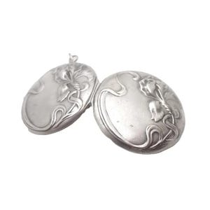 May include: A silver-toned metal locket featuring an embossed floral design with flowing, curved lines. The locket is oval-shaped and consists of two connected pieces, each displaying a matching relief pattern of stylized flowers and stems.