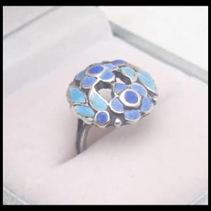 May include: A silver ring with a floral design. The ring features a cluster of blue and light blue enamel petals. The ring is set in a light-coloured box.