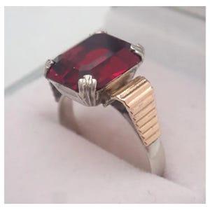 Op de afbeelding: Een zilver- en goudkleurige ring met een grote, rechthoekige, dieprode edelsteen. De ring heeft een tweekleurig ontwerp, met een zilveren band en gouden accenten aan de zijkant van de edelsteen. De edelsteen is het middelpunt.