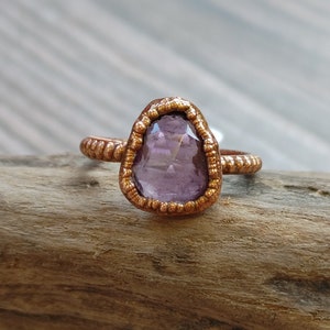 May include: A copper-colored ring with a teardrop-shaped, light purple gemstone. The gemstone is set in a textured bezel, and the band has a twisted rope design. The ring is displayed on a piece of wood.