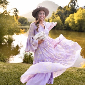May include: A woman wearing a light purple maxi dress with a white crocheted bag and a wide-brimmed hat, standing in a field with a lake in the background.