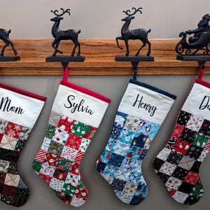 May include: Four Christmas stockings hang from a wooden ledge, each with a name: Mom, Sylvia, Henry, and Dad. The stockings have patchwork designs with festive patterns and are held by black metal stocking holders shaped like reindeer and Santa's sleigh.