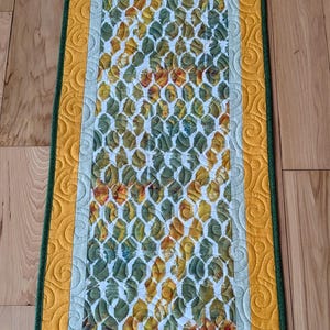 Könnte beinhalten: Rechteckiger, gesteppter Tischläufer mit einem sich wiederholenden Muster aus grünen, gelben und orangefarbenen Formen auf weißem Grund. Der Läufer hat einen gelben Rand mit einem dekorativen Wirbelmuster und einen grünen Rand.