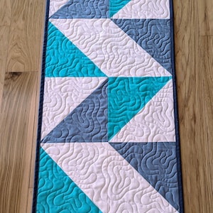 May include: A quilted table runner with a geometric pattern of white, teal, and gray triangles. The runner has a dark blue border.