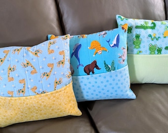 Pode incluir: Três almofadas decorativas com bolsos. As almofadas são azuis com diferentes padrões de animais. A primeira almofada tem um padrão de girafa, a segunda tem um bicho-preguiça, uma baleia e um urso, e a terceira tem um padrão de sapo verde e jacaré.