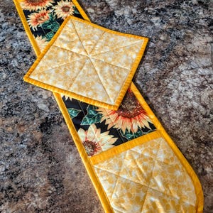 May include: A double oven mitt featuring a sunflower print on a black background with yellow quilted fabric ends and a matching square pot holder. The set is trimmed with bright yellow binding and features a diamond-stitched quilted pattern.