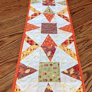 May include: A quilted table runner with a fall theme. The runner is made of various fabrics with autumn colors and patterns, including houses, leaves, and plaid. The runner has an orange border.