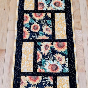 May include: A rectangular table runner featuring a sunflower design. The runner has a black background with square and rectangular panels of sunflowers in orange, yellow, and green. The edges are black with a yellow border.