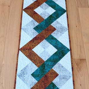 Puede incluir: Camino de mesa acolchado con un diseño geométrico. El camino de mesa presenta formas de diamante que se cruzan en tonos de verde azulado, marrón y gris, sobre un fondo blanco. Los bordes están acabados con un borde marrón, creando un acento decorativo.