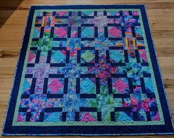 May include: A colorful quilt with a geometric pattern. The quilt features squares of various colors, including blue, pink, green, and purple. Dark blue lines create a grid pattern. The quilt has a dark blue border and a light green inner border.