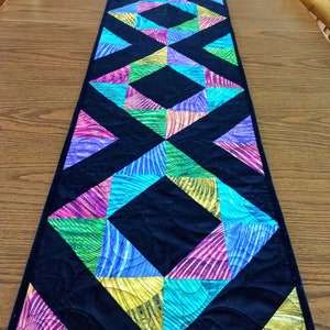 Op de afbeelding: Een zwarte gestikte tafelrunner met een geometrisch patroon van kleurrijke driehoeken. De driehoeken zijn in tinten paars, roze, groen, blauw en geel.