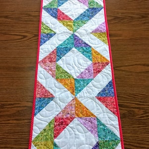 May include: A colourful table runner with a geometric pattern of triangles and squares. The runner is made of fabric with a floral print and has a white background. The runner has a red border.
