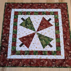 May include: A square Christmas quilt featuring a central design of four stylized Christmas trees. The trees alternate between green and red, set against a white background with gold floral accents. The quilt has a red border with a floral pattern.