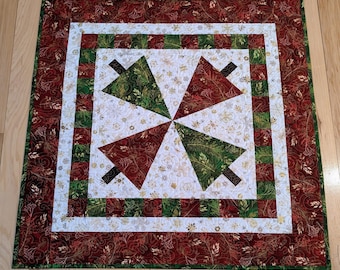 May include: A square Christmas quilt featuring a central design of four stylized Christmas trees. The trees alternate between green and red, set against a white background with gold floral accents. The quilt has a red border with a floral pattern.