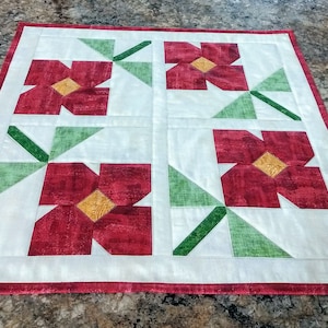 May include: A white table runner with a red border and four red flowers with yellow centers. The flowers are surrounded by green leaves.