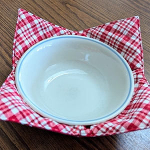 May include: A red and white checkered bowl cozy with a white bowl inside. The cozy is made of fabric and has a star shape.